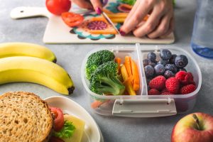 A picture of someone packing a lunch - cutting up raw peppers and tomatoes and putting them into a container along with raw broccoli and berries. Also pictured is a sandwich on whole grain bread, an apple and banana with a bottle of water.