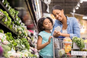 A picture of a parent and child grocery shopping. They are in the produce section with many vegetables pictured. They are reading a grocery list.